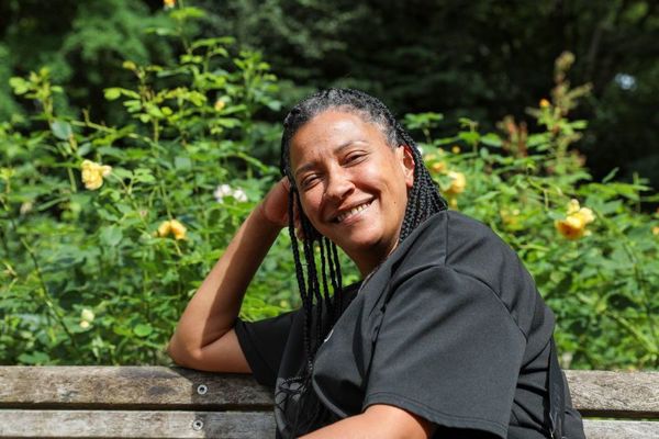 A joyful woman with braided hair sits on a park bench, smiling at the camera, with sunlit trees and yellow flowers in the background.