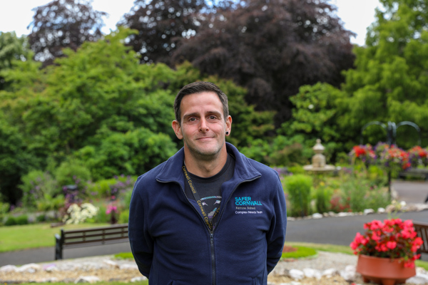 A man wearing a blue zip-up sweatshirt with "cornwall" embroidered on it is standing in a garden with trees, flowers, and a bench in the background.